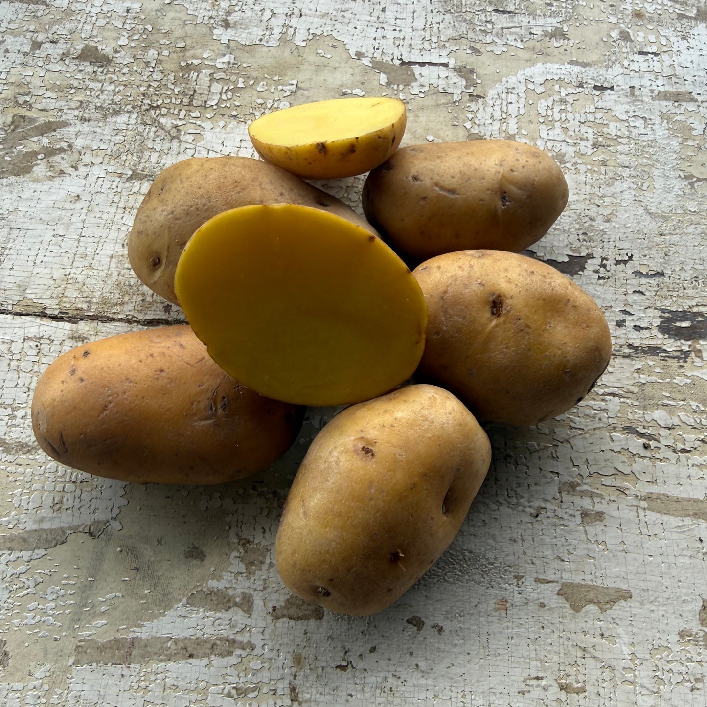 A cluster of whole and halved yellow potatoes on a distressed white wooden surface, showing their tan skin and bright yellow-orange flesh.